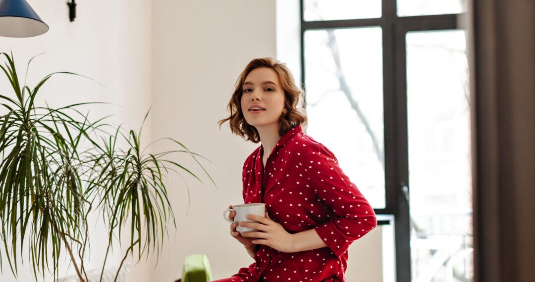 Appealing european girl in pajama holding cup of coffee and looking at camera. Indoor shot of young woman posing in morning at home.